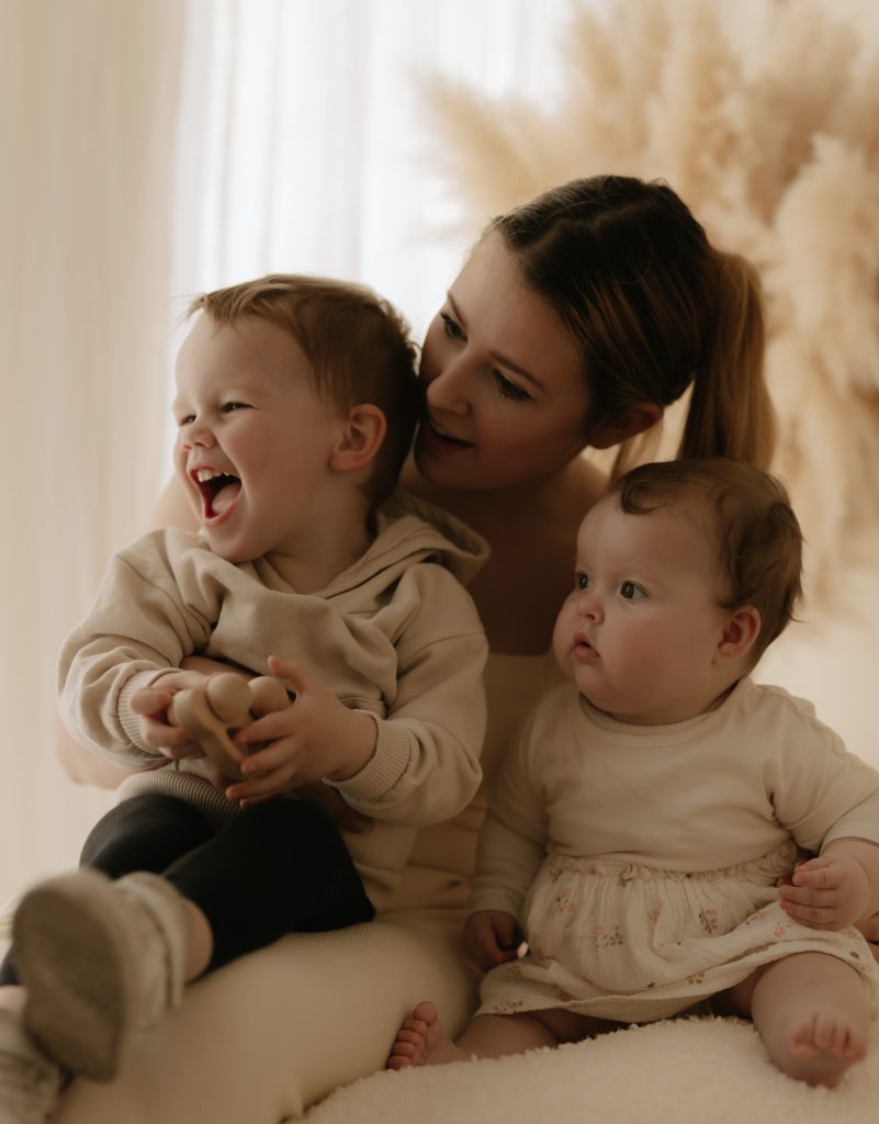 Mum having photoshoot with her toddler and baby girl sat on her lap smiling.